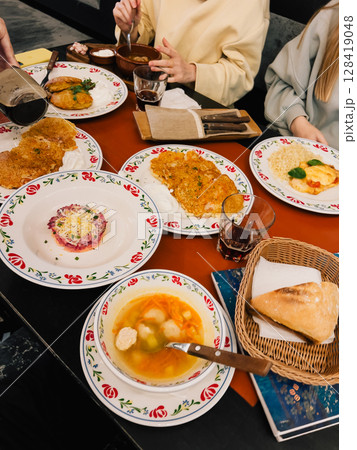 People are sharing a meal of potato pancakes, soup, salad, and bread, served on decorative plates and bowls in a cozy restaurant setting, highlighting the rich culinary traditions of Eastern Europe People are sharing a meal of potato pancakes, soup, salad, and bread, served on decorative plates and bowls in a cozy restaurant setting, highlighting the rich culinary traditions of Eastern Europe 128419048