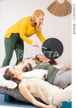 Group therapy session with Tibetan drum for group of women lying on wooden floor surrounded by copper bowls. Meditation and relaxation 128419072