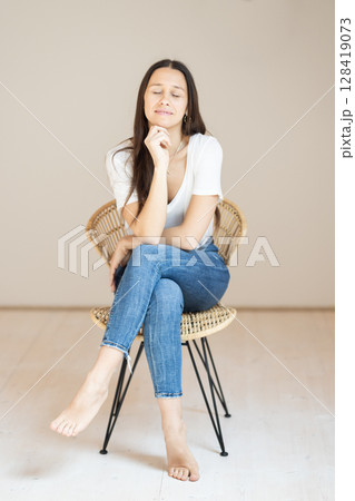 Portrait of confident beautiful woman with long brown hair, wearing casual clothes, sitting on chair in tight jeans and white t-shirt, studio background Portrait of confident beautiful woman with long brown hair, wearing casual clothes, sitting on chair in tight jeans and white t-shirt, studio background 128419073