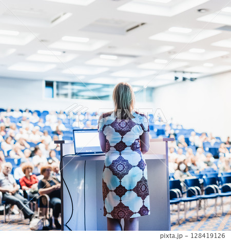 Female speaker giving a talk on corporate business conference. Unrecognizable people in audience at conference hall. Business and Entrepreneurship event. Female speaker giving a talk on corporate business conference. Unrecognizable people in audience at conference hall. Business and Entrepreneurship event. 128419126