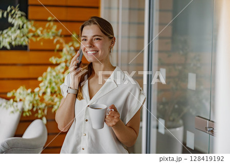 A Cheerful Young Woman Engaged in a Delightful Conversation on the Phone While Holding Her Coffee 128419192
