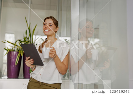 A young woman in a modern office holds a tablet and coffee, looking professional and thoughtful A young woman in a modern office holds a tablet and coffee, looking professional and thoughtful 128419242