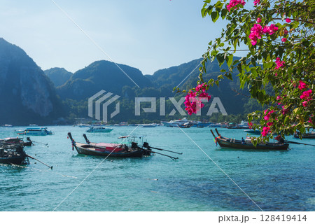 Serene coastal scene in Koh Phi Phi, Thailand, with turquoise waters, longtail boats, limestone cliffs, and vibrant pink bougainvillea flowers. Serene coastal scene in Koh Phi Phi, Thailand, with turquoise waters, longtail boats, limestone cliffs, and vibrant pink bougainvillea flowers. 128419414