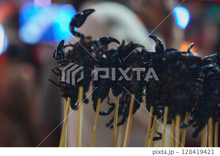 Cooked skewered scorpions showcased in a lively Thai market with blurred colorful lights and figures, suggesting a bustling festive atmosphere. Cooked skewered scorpions showcased in a lively Thai market with blurred colorful lights and figures, suggesting a bustling festive atmosphere. 128419421
