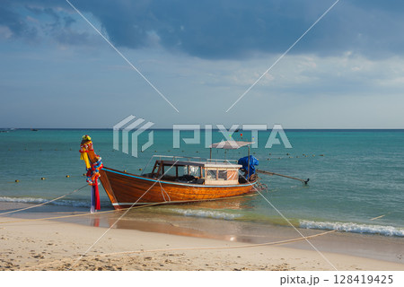 A longtail boat adorned with colorful ribbons rests on a sandy beach in Thailand. Calm turquoise waters and buoys are visible under a partly cloudy sky. 128419425