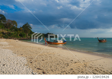 A tranquil beach scene in Thailand featuring turquoise waters, two ribbon adorned longtail boats, sandy shores, lush greenery, and a partly cloudy sky. 128419426