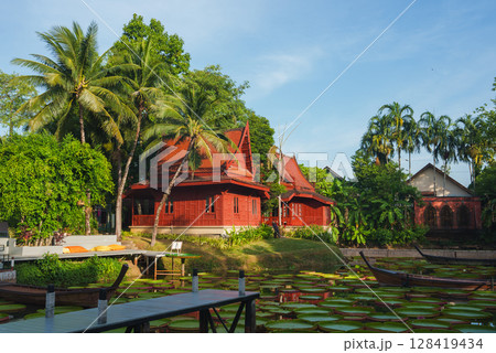 Thai style red wooden buildings with steep roofs surrounded by greenery, a pond with lily pads and boats, and a wooden dock under a blue sky. Thai style red wooden buildings with steep roofs surrounded by greenery, a pond with lily pads and boats, and a wooden dock under a blue sky. 128419434