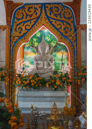 Seated white Buddha statue in a gold adorned shrine, framed by blue patterns, with a mural of trees and mountains, surrounded by offerings in Thailand. Seated white Buddha statue in a gold adorned shrine, framed by blue patterns, with a mural of trees and mountains, surrounded by offerings in Thailand. 128419439