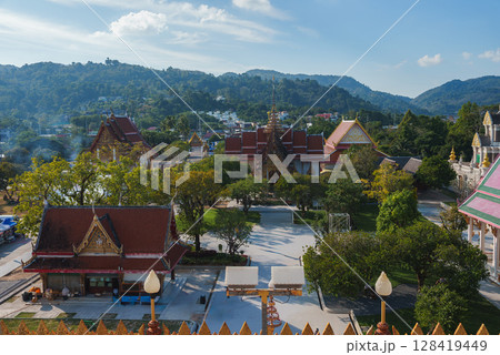 Wat Chalong in Phuket, Thailand, features red and gold roofs, lush greenery, mountain views, and intricate temple details under a clear blue sky. Wat Chalong in Phuket, Thailand, features red and gold roofs, lush greenery, mountain views, and intricate temple details under a clear blue sky. 128419449