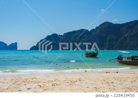 Golden sand, turquoise waters, and anchored longtail boats with limestone cliffs in the background under a bright blue sky on Koh Phi Phi islands. 128419450