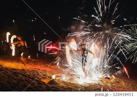 Performers spin fire props, creating light trails, as fireworks burst above a sandy beach. A dark background highlights the fiery spectacle and audience. 128419451
