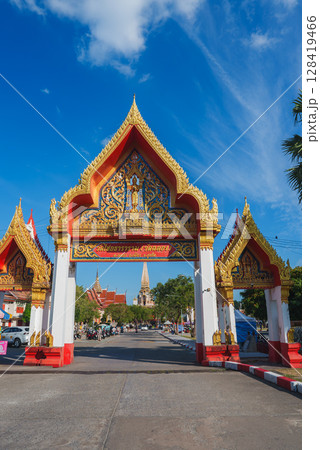 The intricately carved golden and red gate of Wat Chalong in Phuket, Thailand, with a temple spire, blue sky, greenery, and distant visitors visible. 128419466