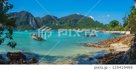 Turquoise waters, lush green cliffs, and anchored boats define this serene Koh Phi Phi beach scene, framed by vibrant foliage and a clear blue sky. 128419469