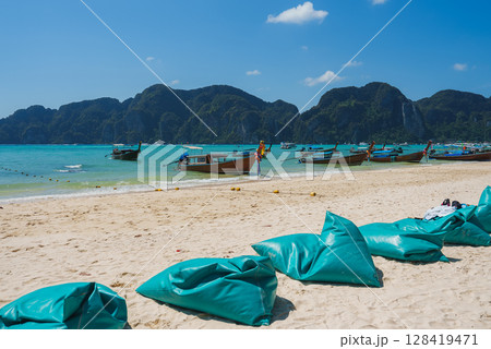 A sandy beach on Koh Phi Phi, Thailand, featuring turquoise waters, limestone cliffs, longtail boats, teal bean bags, and a clear blue sky. 128419471