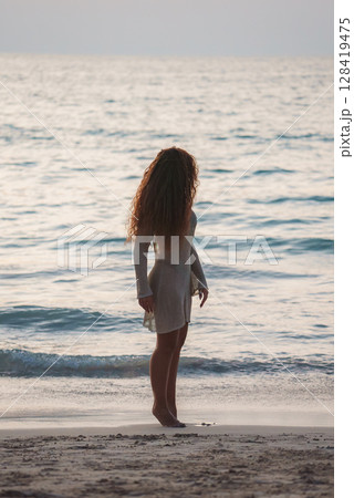 A woman with long curly hair stands barefoot on a sandy beach, wearing a light dress. She faces the calm ocean under soft, warm lighting in Phuket or Koh Phi Phi. 128419475