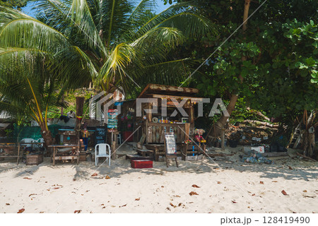 A wooden beachside shack with colorful signs and a menu board sits under lush palm trees, surrounded by chairs and tables on a sandy beach. A wooden beachside shack with colorful signs and a menu board sits under lush palm trees, surrounded by chairs and tables on a sandy beach. 128419490