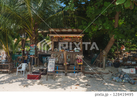 A wooden beachside shack under palm trees, featuring colorful garlands, handmade signs, and a casual setup with drinks and snacks on display. A wooden beachside shack under palm trees, featuring colorful garlands, handmade signs, and a casual setup with drinks and snacks on display. 128419495