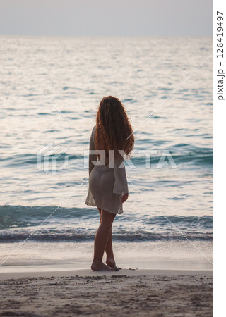 A barefoot woman with long curly hair stands on a sandy beach, facing the calm ocean. Soft lighting suggests sunrise or sunset, with gentle waves at the shore. 128419497