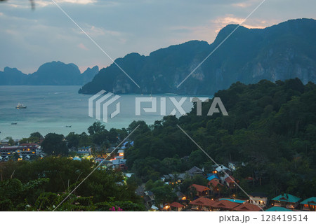 Lush green hills, a village with colorful rooftops, turquoise waters, boats, and limestone cliffs under soft dawn or dusk lighting in Koh Phi Phi, Thailand. Lush green hills, a village with colorful rooftops, turquoise waters, boats, and limestone cliffs under soft dawn or dusk lighting in Koh Phi Phi, Thailand. 128419514