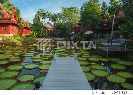 A tranquil pond with green lily pads, a white pier, and a small boat, surrounded by Thai style red wooden buildings and lush tropical greenery. A tranquil pond with green lily pads, a white pier, and a small boat, surrounded by Thai style red wooden buildings and lush tropical greenery. 128419522