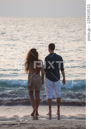 A couple faces the ocean on a sandy beach in Thailand, with soft warm light, gentle waves, and a calm horizon blending with the sea. A couple faces the ocean on a sandy beach in Thailand, with soft warm light, gentle waves, and a calm horizon blending with the sea. 128419523