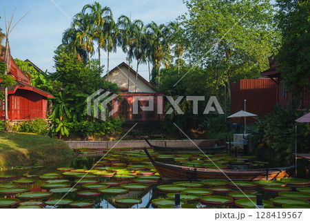 Serene scene in Thailand with red wooden buildings, a temple, lush greenery, palm trees, a calm waterway with lily pads, and a wooden boat. Serene scene in Thailand with red wooden buildings, a temple, lush greenery, palm trees, a calm waterway with lily pads, and a wooden boat. 128419567