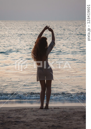 A woman in a light dress stands on a beach at sunset, arms raised gracefully. Calm waves and golden light reflect off the ocean in Thailand. A woman in a light dress stands on a beach at sunset, arms raised gracefully. Calm waves and golden light reflect off the ocean in Thailand. 128419601
