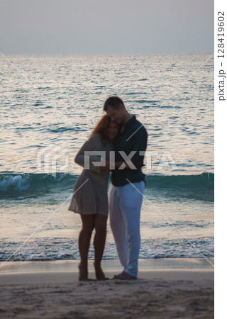 A man in a dark shirt and white pants and a woman in a light dress stand close on a sandy beach at sunset, with gentle waves in the background. A man in a dark shirt and white pants and a woman in a light dress stand close on a sandy beach at sunset, with gentle waves in the background. 128419602
