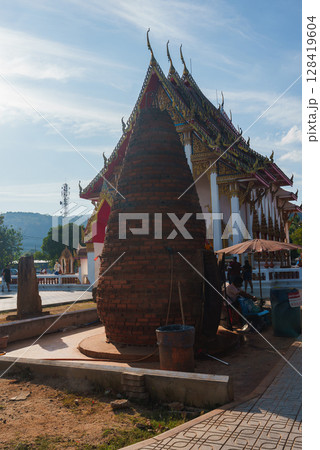 A traditional Thai temple in Phuket, Thailand, featuring gold accented roofs, a brick structure in the foreground, and a backdrop of hills and trees. A traditional Thai temple in Phuket, Thailand, featuring gold accented roofs, a brick structure in the foreground, and a backdrop of hills and trees. 128419604