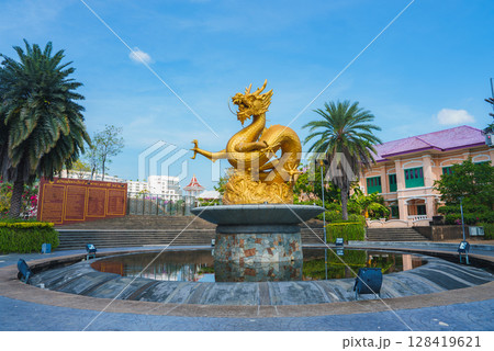 Golden dragon statue atop a circular fountain surrounded by gardens, palm trees, and a pink roofed building under a clear blue sky in Thailand. Golden dragon statue atop a circular fountain surrounded by gardens, palm trees, and a pink roofed building under a clear blue sky in Thailand. 128419621