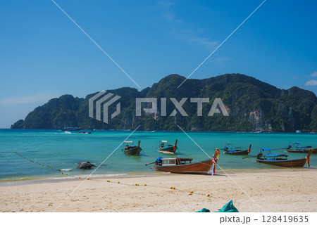 Turquoise waters, limestone cliffs, and anchored longtail boats with colorful ribbons on a sandy beach under a clear blue sky in Koh Phi Phi, Thailand. 128419635