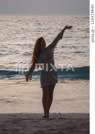 A person in a light dress stands on a beach at sunset, arm raised in a dynamic pose. Ocean waves and golden light create a serene silhouette effect. 128419640