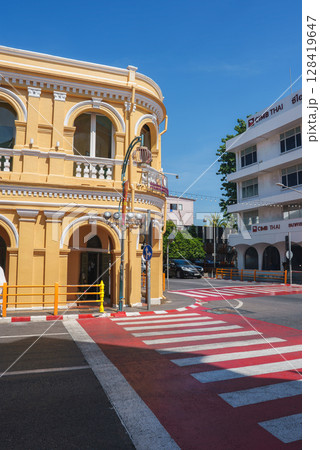 A vibrant street in Phuket, Thailand, featuring a yellow Sino Portuguese colonial building, a red and white pedestrian crossing, and a modern white structure. 128419647