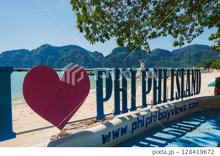 Koh Phi Phi Island beachfront with I Love Phi Phi Island sign, turquoise waters, limestone cliffs, sandy beach, and overhanging tree branches. 128419672