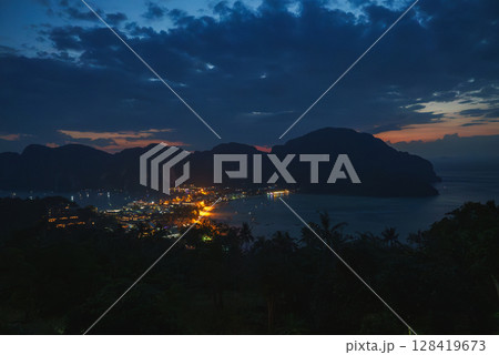 Koh Phi Phi at night with town lights reflecting on calm bay waters, lush vegetation in the foreground, and limestone cliffs in the background. 128419673