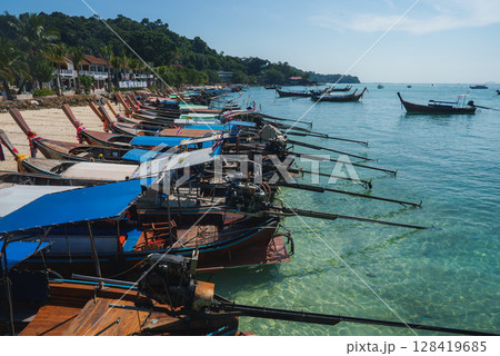 Row of long tail boats with colorful ribbons docked on a beach, turquoise water, green hills, palm trees, and beachfront buildings in the background. Row of long tail boats with colorful ribbons docked on a beach, turquoise water, green hills, palm trees, and beachfront buildings in the background. 128419685