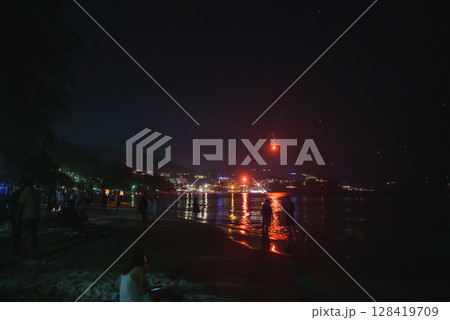 A lively beach in Phuket, Thailand, at night with city lights reflecting on the water, a red lantern in the sky, and silhouettes of people along the shore. 128419709