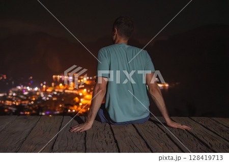 A man in a light blue shirt sits on a wooden platform at night, gazing at a brightly lit cityscape below, surrounded by dark hills in Thailand. 128419713