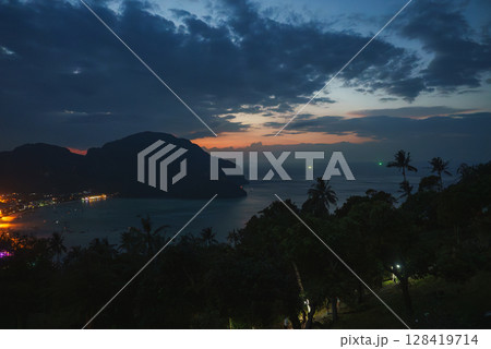 Serene twilight scene of Koh Phi Phi, Thailand, featuring palm silhouettes, glowing coastal lights, calm waters, and distant fishing boat lights. 128419714