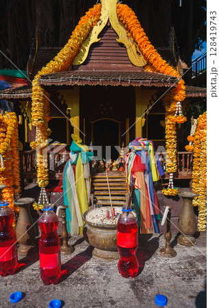 A small Thai spirit house adorned with marigold garlands, colorful fabric, and offerings like red liquid bottles, incense, and figurines under bright sunlight. A small Thai spirit house adorned with marigold garlands, colorful fabric, and offerings like red liquid bottles, incense, and figurines under bright sunlight. 128419743
