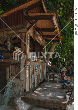 A weathered wooden shack with a corrugated metal roof surrounded by lush greenery, featuring a small counter with items and a seated person nearby. A weathered wooden shack with a corrugated metal roof surrounded by lush greenery, featuring a small counter with items and a seated person nearby. 128419749