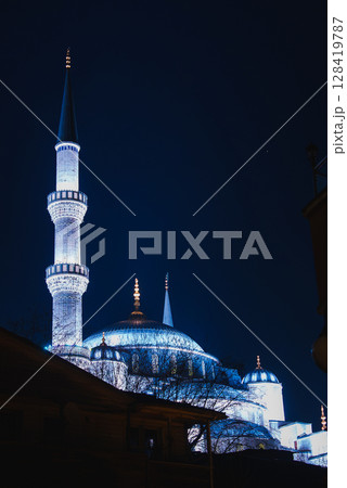 The Sultan Ahmed Mosque, or Blue Mosque, glows with blue and white lights at night. Domes, minaret, and tree silhouettes frame the dark sky. 128419787