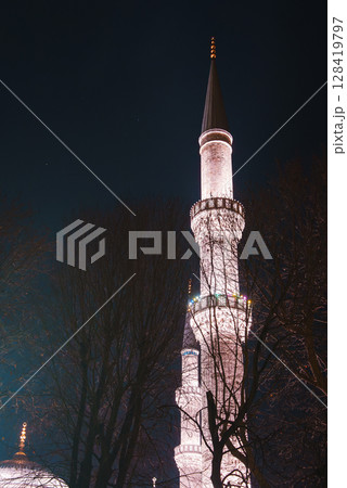 A brightly lit minaret with intricate details and a golden finial stands against a dark night sky, partially framed by bare tree branches in Istanbul, Turkey. 128419797