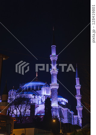 The Sultan Ahmed Mosque, with its domes and six minarets lit in purple and blue, stands against a dark sky, framed by trees and nearby structures. 128419801