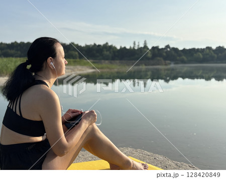 Adult sporty woman relaxing on a yellow exercise mat, listening to music in headphones after workout by lake in forest. Outdoor fitness, recovery, peaceful summer nature scene 128420849