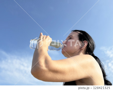 Athletic woman drinking water from bottle against blue sky. Outdoor fitness, hydration, body and self care 128421325