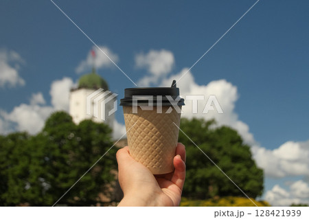Brown paper cup of drink in women's hand on against backdrop of island Olaf's Tower in town Vyborg in blur, Russia on sunny day Brown paper cup of drink in women's hand on against backdrop of island Olaf's Tower in town Vyborg in blur, Russia on sunny day 128421939