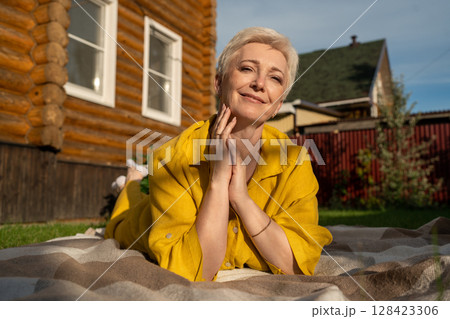 A serene scene of a mature woman dressed in yellow, leisurely relaxing on a blanket in her garden near house, enjoying sun A serene scene of a mature woman dressed in yellow, leisurely relaxing on a blanket in her garden near house, enjoying sun 128423306