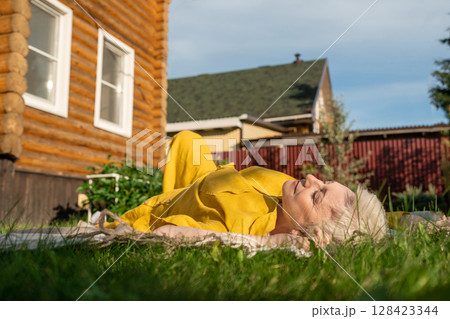 A serene scene of a mature woman dressed in yellow, leisurely relaxing on a blanket in her garden near house, enjoying sun A serene scene of a mature woman dressed in yellow, leisurely relaxing on a blanket in her garden near house, enjoying sun 128423344