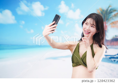 快晴のビーチでスマホで自撮りをする水着を着た若い女性 128423362